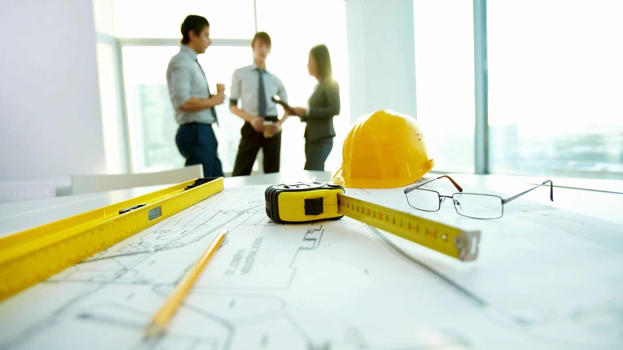 Three project managers discuss planning on the construction jobsite with blue prints, hard hats and a tape measure on the table.