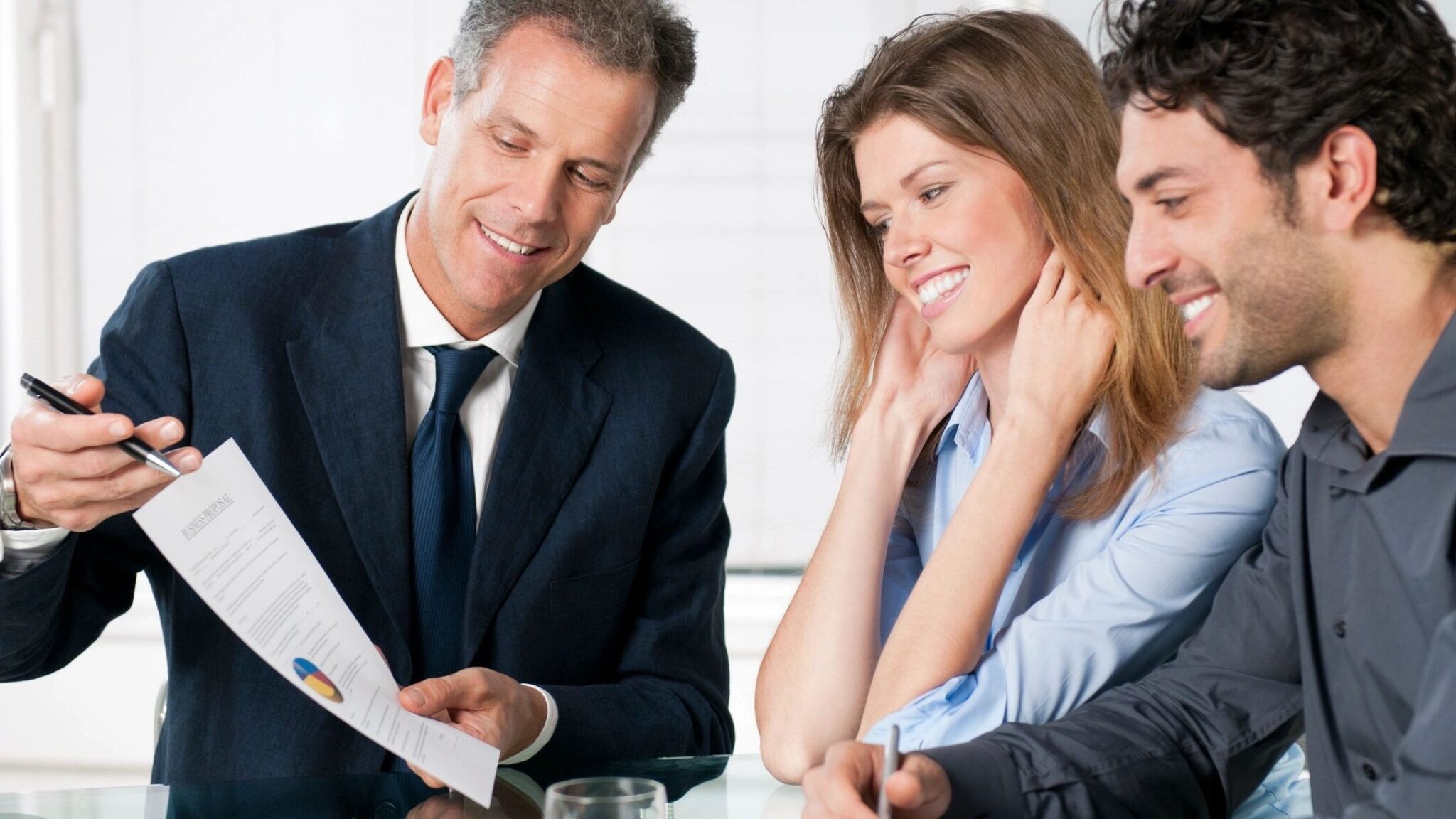 An agent smiles as he shows his two clients paperwork at a table. The clients are also smiling.