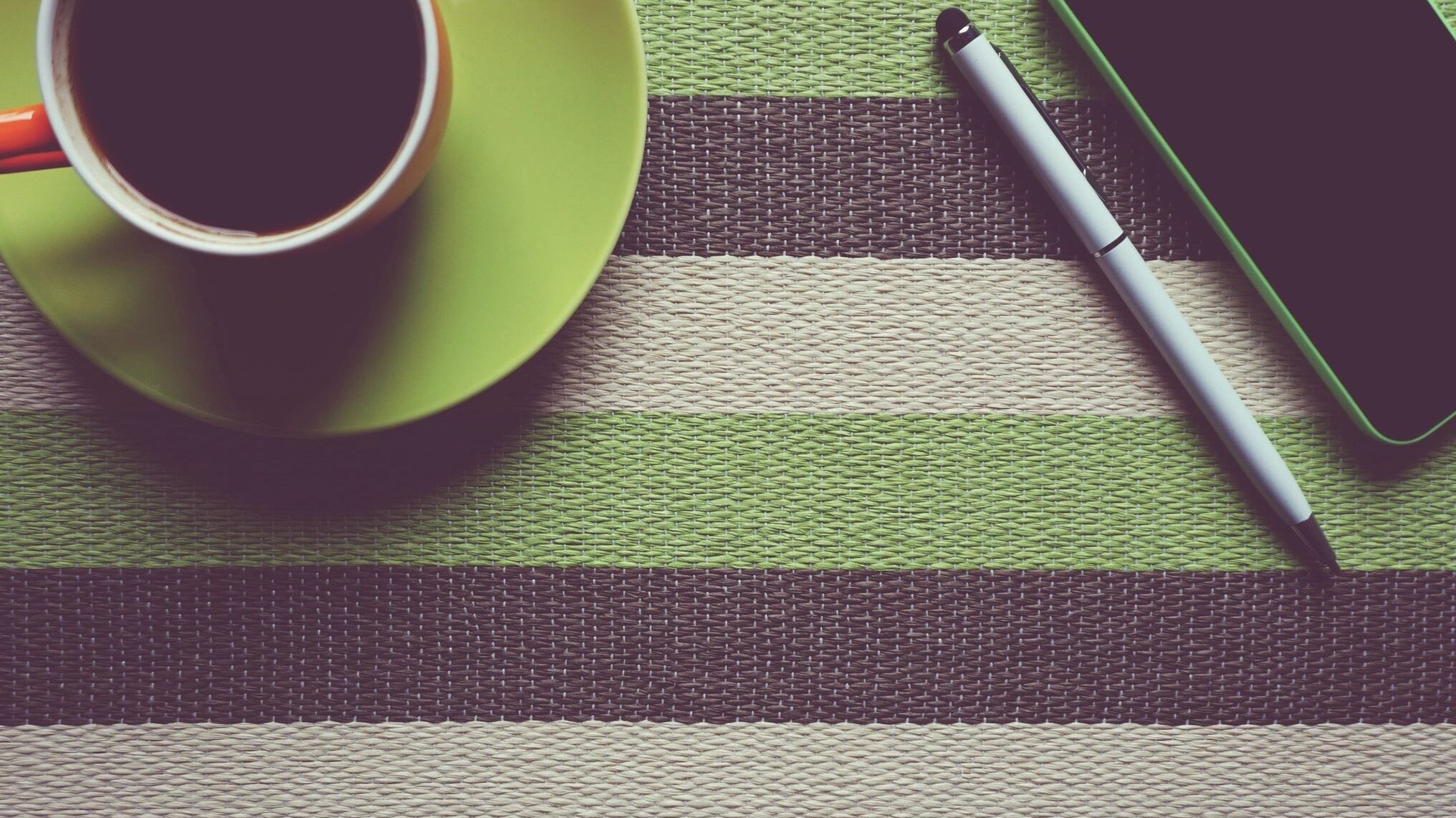 A green and black stripped placemat matches the saucer, coffee cup, pen and cell phone, that sits on it.