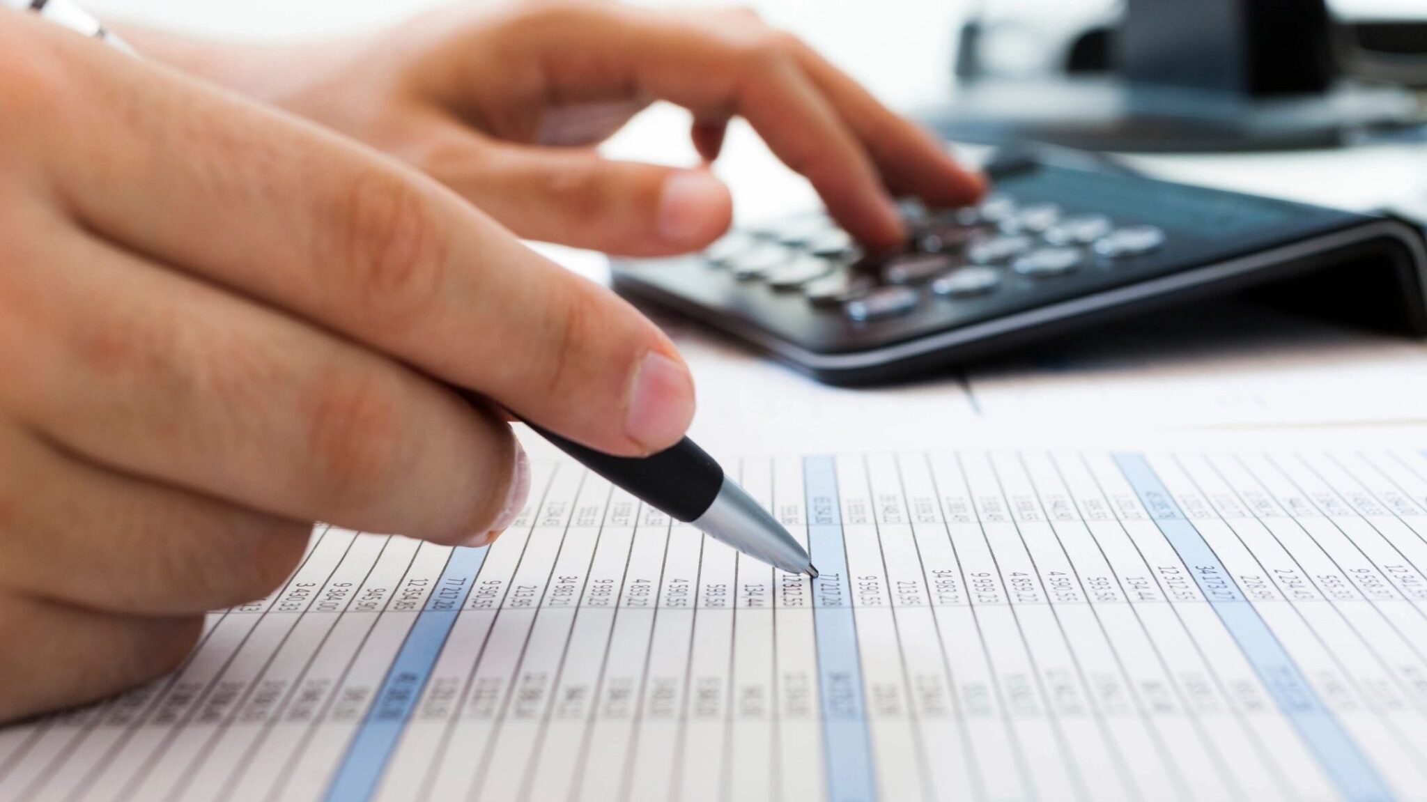 A person points to figures in a financial report at their desk as they key-in figures into a calculator with their left hand.